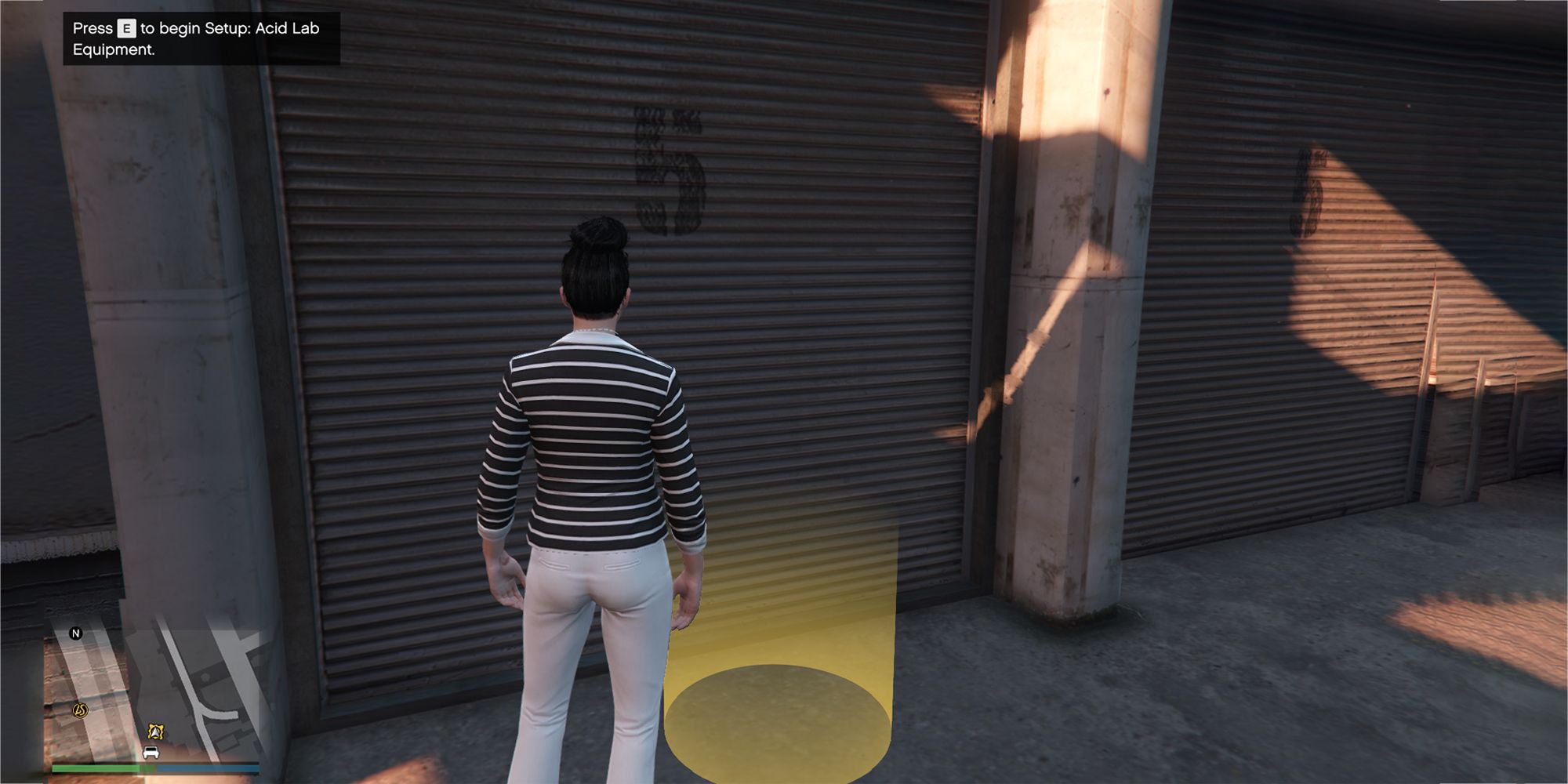 A imagem mostra um jogador do Grand Theft Auto Online do lado de fora de um armazém próximo a um marcador de círculo amarelo no chão.  O texto no canto superior esquerdo diz 'Pressione E para iniciar a configuração do equipamento de laboratório de ácido'.