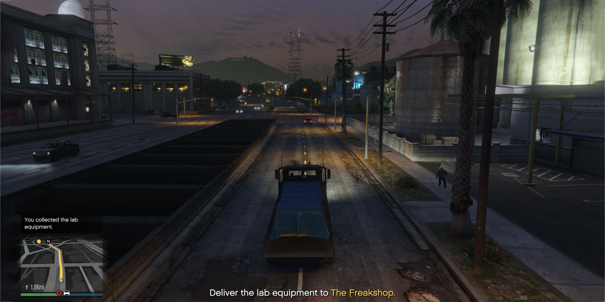 A imagem mostra um jogador do Grand Theft Auto Online em um caminhão com uma carroceria coberta por um lençol azul na estrada principal.  O texto na parte inferior central diz 'Entregue o equipamento de laboratório para The Freakshop'.